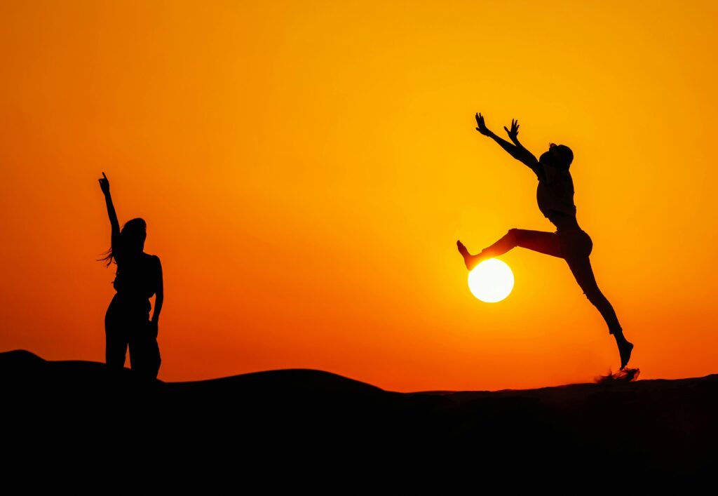 Two silhouetted people jumping joyfully during a vibrant sunset in Dubai's desert.
